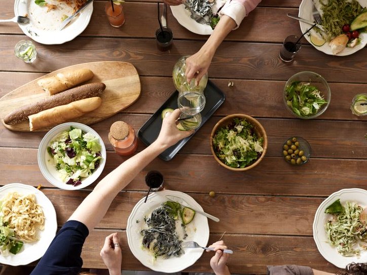 A family table seen from above: shared plates of pasta and salad, bread on a board, hands reaching across.